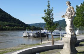 Statue am Donauufer mit Anlegestelle und bewaldeten Hügeln im Hintergrund., © Donau NÖ Tourismus JMZ