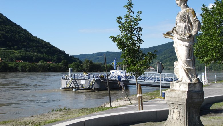 Statue am Donauufer mit Fähranleger und bewaldeten Hügeln im Hintergrund., © Donau NÖ Tourismus/JMZ