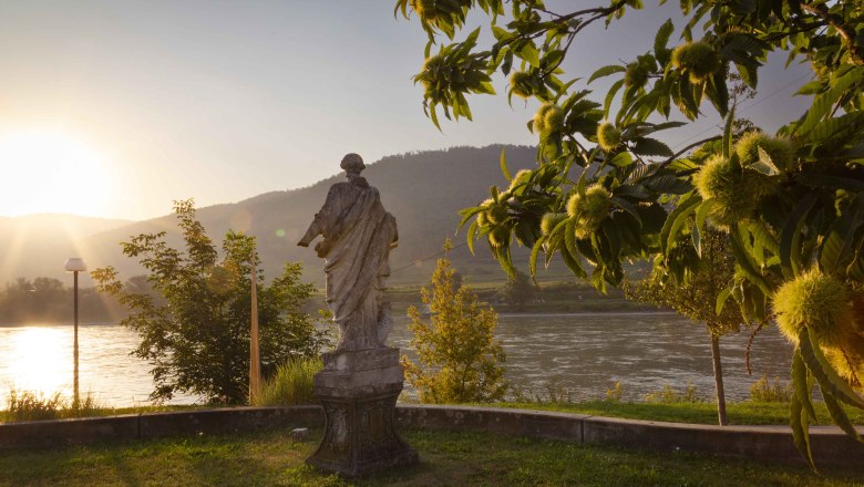 Statue am Flussufer bei Sonnenuntergang, umgeben von Kastanienbäumen., © Donau Niederoesterreich_Barbara Elser