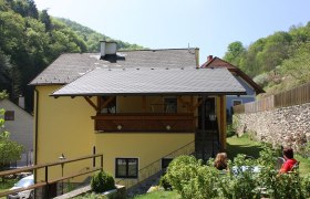 Gelbes Haus mit Balkon, umgeben von grüner Landschaft und Steinmauer., © Bergkirchner