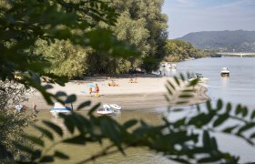 Sandstrand an einem Fluss mit Booten und Menschen, umgeben von Bäumen., © Donau NÖ/Barbara Elser